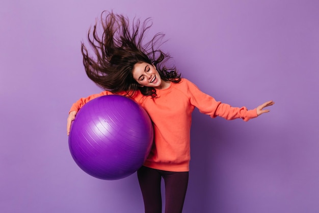 Привлекательная женщина в оранжевом спортивном свитере и черных леггинсах танцует и держит фитбол на фиолетовом фоне