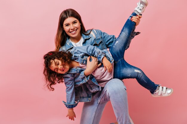 Attractive woman playing with little daughter on pink background. Studio shot of mom and preteen kid in denim jackets.