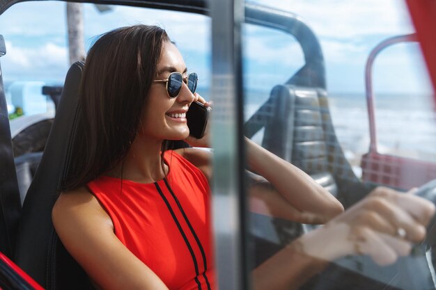 海岸近くで車を運転し、ビーチで彼女を待っている友人に電話をかけている赤いビキニの魅力的な女性セクシーな女性モデルの笑顔は携帯電話を使用して島のリゾートを横切ってジープを運転します