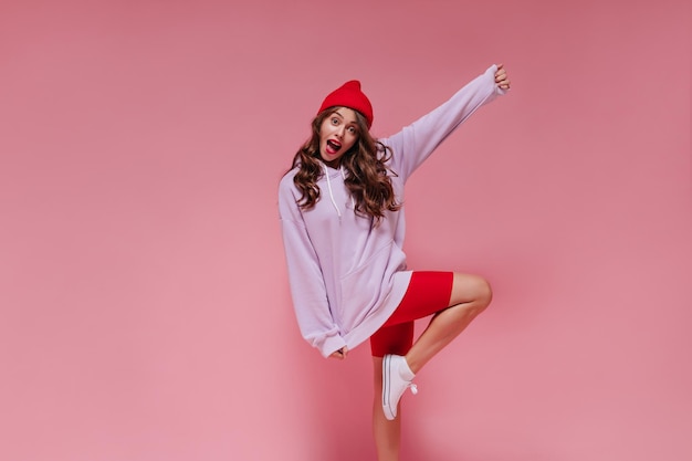 Attractive woman in red hat cycling shorts and purple hoodie moves on pink background Curly girl rises leg on isolated backdrop