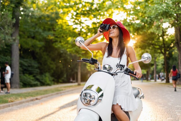 거리에서 오토바이를 타는 매력적인 여자, 여름 휴가 스타일, 여행, 미소, 행복, 재미, 세련된 복장, 모험, 빈티지 사진 카메라로 사진 촬영