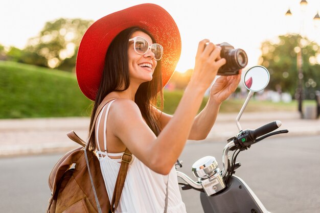 Attractive woman riding on motorbike in street, summer vacation style, traveling, smiling, having fun, stylish outfit, adventures, taking pictures on vintage photo camera, wearing leather backpack