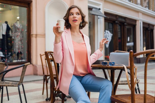 Attractive woman in romatic mood smiling in happiness sitting at table wearing pink jacket, stylish apparel, waiting for boyfriend on a date in cafe, drinking cappuccino, exited face expression