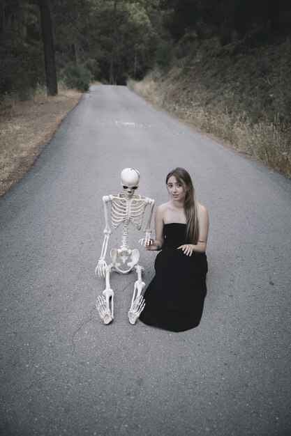 Attractive woman sitting on road with decorative skeleton of human and looking at camera