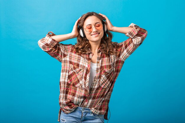 Attractive woman smiling enjoying listening to music in headphones in checkered shirt and jeans isolated on blue studio background, wearing pink sunglasses