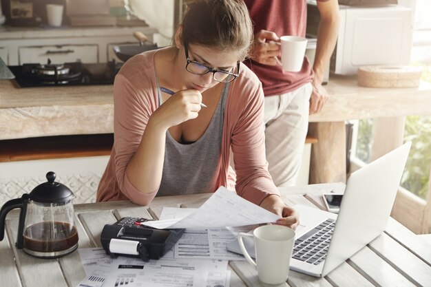 Привлекательная женщина в очках с серьезным и сосредоточенным взглядом держит ручку, заполняя бумаги, подсчитывая счета, сокращая семейные расходы, пытаясь сэкономить деньги, чтобы совершить крупную покупку