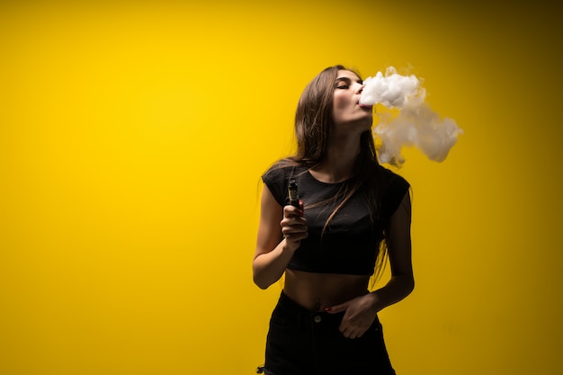 Attractive woman standing and vaping on yellow wall.