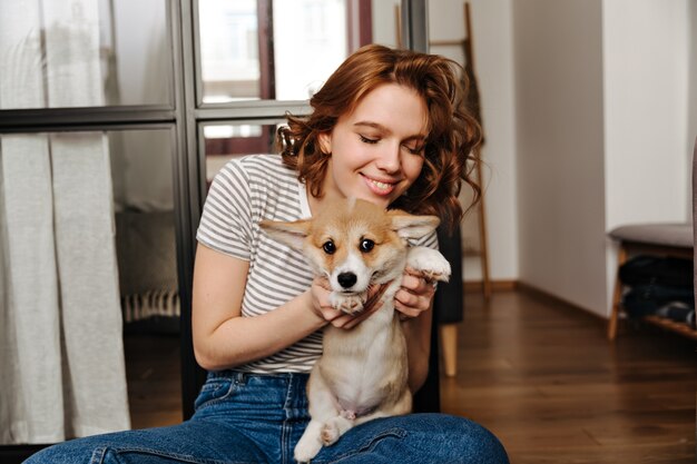 Attractive woman in striped T-shirt plays with corgi in her apartment.