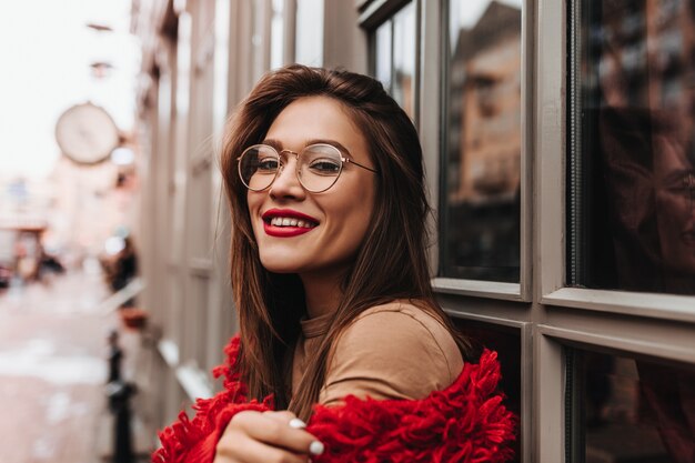 Attractive woman in stylish red outfit posing on street. Dark-haired woman with bright lipstick is smiling.