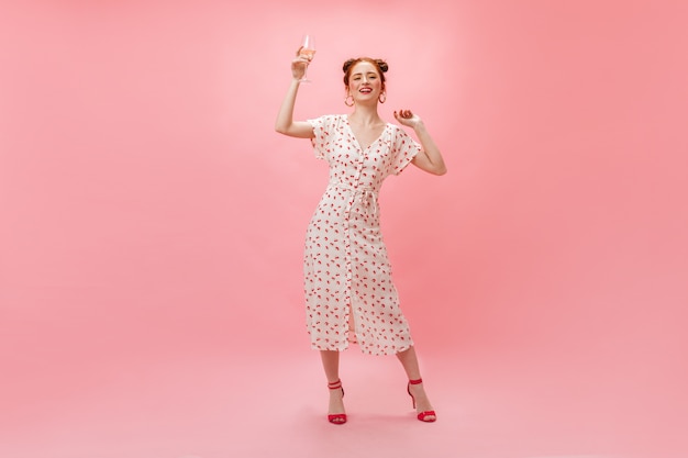 Attractive woman in stylish white polka-dot dress dancing with glass of champagne on pink background.