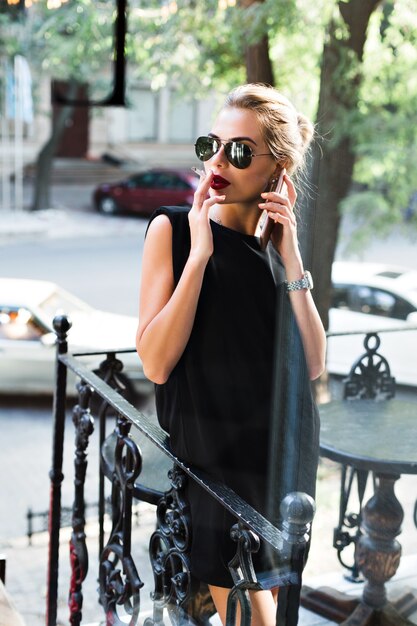 Attractive woman in sunglasses in black short dress smoking cigarette on terrace. She is speaking on phone, looking to side.