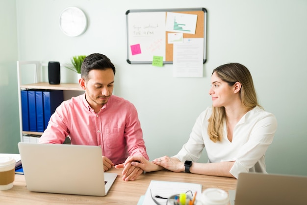 Attractive woman touching the hand of her male coworker and flirting at the office. Latin man feeling uncomfortable for the inappropriate behavior of his business partner