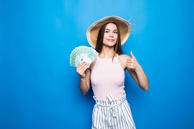 Привлекательная женщина носит в соломенной шляпе, показывая банкноты в 100 долларов, большой палец вверх, изолированные над синей стеной.