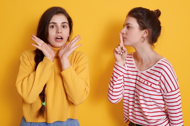 Attractive woman wearing striped shirt keeps finger near lips while telling secret to her friend