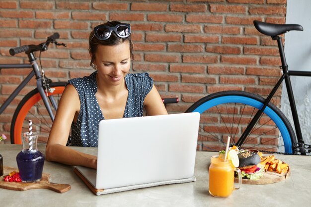 점심 시간 동안 열린 노트북 컴퓨터 앞에 앉아 고속 인터넷 연결을 사용하여 온라인 쇼핑을하는 그녀의 머리에 선글라스를 착용 한 매력적인 여자
