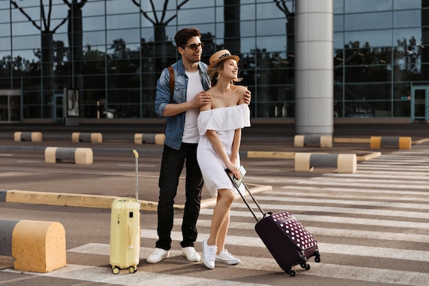 Attractive woman in white dress hat holds suitcase Brunette man in denim jacket black pants and sunglasses hugs girlfriend near airport