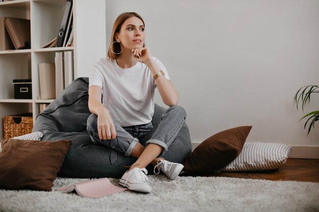 Attractive woman with beautiful massive earrings and in white T-shirt with smile, posing while sitting in armchair.