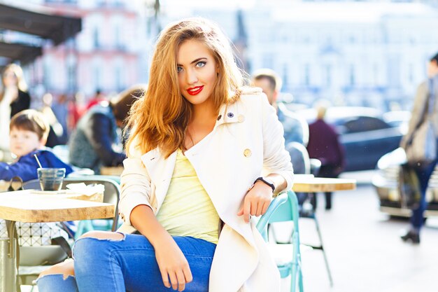 Привлекательная женщина со светлыми волосами, сексуальная улыбающаяся девушка в открытом кафе, фоне города. В джинсах с дырочками, в белом халате.