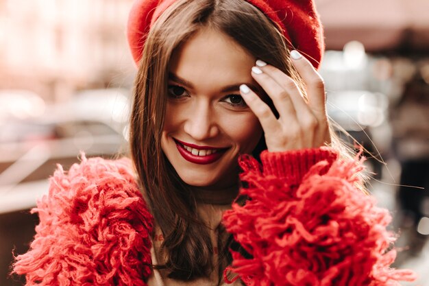 Attractive woman with bright lips smiling and touching her dark hair. Closeup portrait of lady in red outfit and headdress.