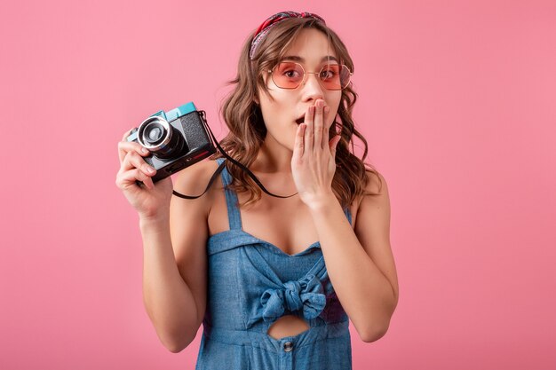 Attractive woman with funny surprised emotional face expression with vintage camera in denim dress and sunglasses on pink background