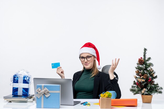 Attractive woman with her santa claus hat and wearing eyeglasses sitting at a table and holding bank card asking something in the office
