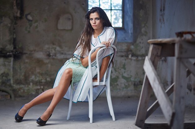 Attractive woman with long dark hair in white dress posing on chairwith window behind her.