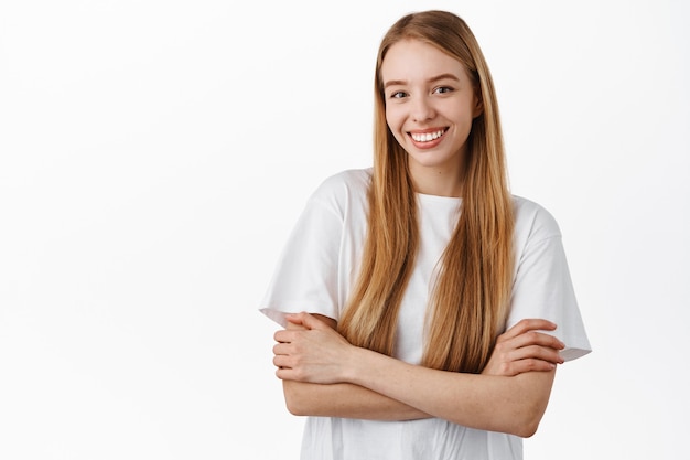 Free photo attractive woman with long straight blond hair, cross arms on chest, looking confident and determined, smiling happy, standing in t-shirt against white wall