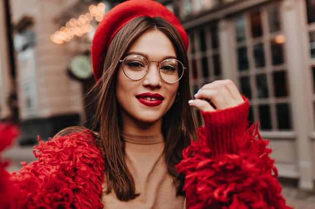 Attractive woman with red lips makes selfie on street. Shot of brunette in glasses dressed in stylish hat, red jacket and beige top.