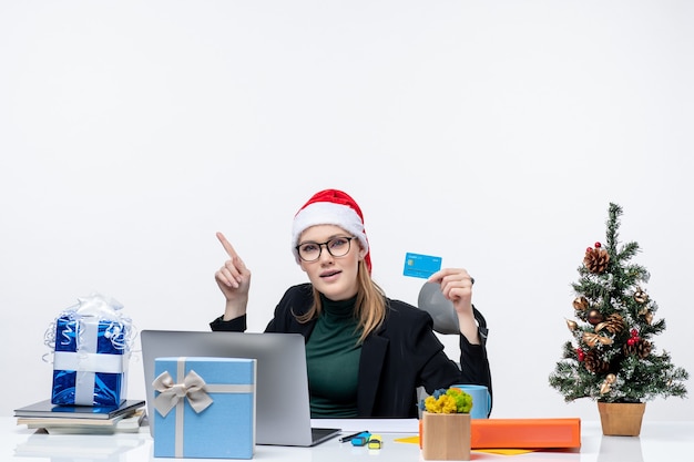 サンタクロースの帽子をかぶって、テーブルのクリスマスプレゼントに座って、オフィスで上を見ている銀行カードを保持している眼鏡をかけている魅力的な女性