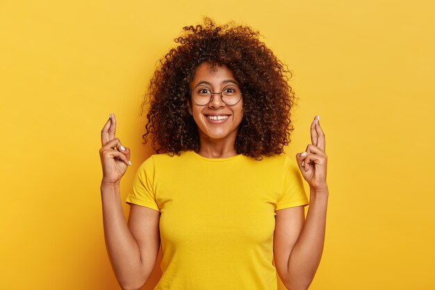 Attractive woman in yellow t shirt, crosses fingers, hopes in lucky future, smiles pleasantly, poses against vivid background, prays indoor