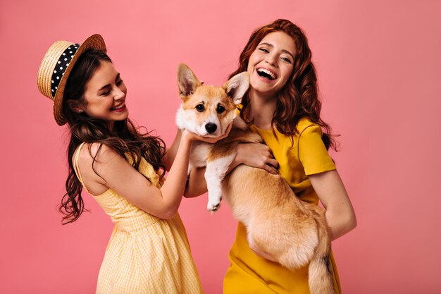 ピンクの背景にカメラのポーズをとってかわいいコーギーと犬の巻き毛の若いブルネットを保持している黄色のドレスの魅力的な女性