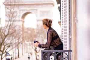 無料写真 パジャマ姿の魅力的な陽の女性は、パリの街で朝のバルコニーでコーヒーを飲んでいます。凱旋門の眺め。