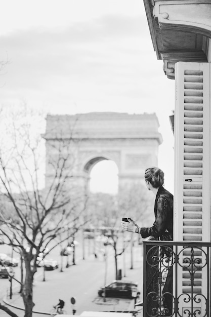 Foto gratuita attraente donna yang in pigiama sta bevendo caffè sul balcone al mattino nella città di parigi. veduta dell'arco trionfale.