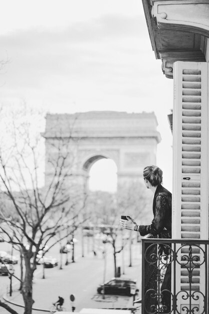 Привлекательная женщина-ян в пижаме пьет кофе на балконе утром в Париже. вид на триумфальную арку.