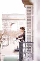 Free photo attractive  yang woman in pajama is drinking coffee on balcony in the morning in city paris. view of the triumphal arch.