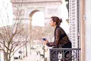 Foto gratuita attraente donna yang in pigiama sta bevendo caffè sul balcone al mattino nella città di parigi. veduta dell'arco trionfale.