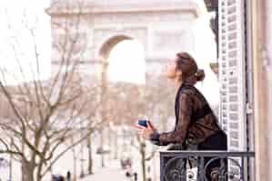 Foto gratuita attraente donna yang in pigiama sta bevendo caffè sul balcone al mattino nella città di parigi. veduta dell'arco trionfale.