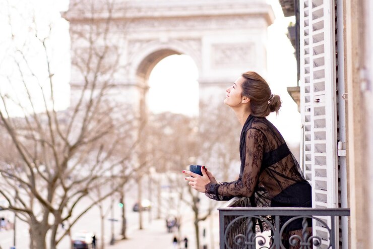 パジャマ姿の魅力的な陽の女性は、パリの街で朝のバルコニーでコーヒーを飲んでいます。凱旋門の眺め。
