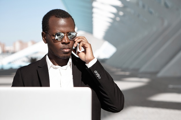 Привлекательный молодой афроамериканец бизнесмен носить стильные солнцезащитные очки и формальную одежду, сидя в городском кафе перед ноутбуком, имея телефонный разговор с партнерами во время ожидания кофе