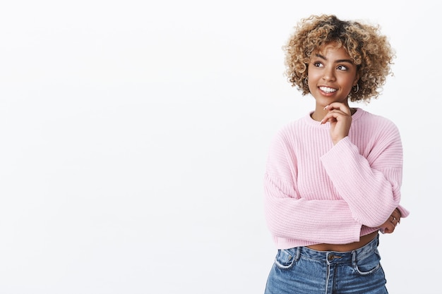 Free photo attractive young african-american female student with blond hair holding hand on chin and looking delighted at upper left corner as thinking, posing thoughtful pleased with idea came to mind