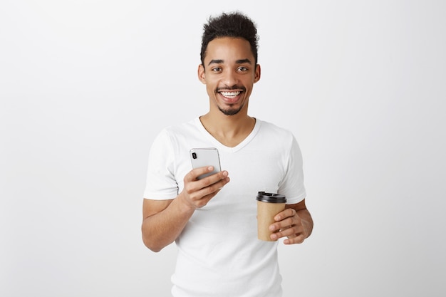 Attractive young african american man looking pleased, holding mobile phone and drinking takeaway coffee