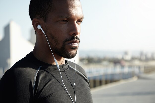 Attractive young African American runner or jogger dressed in black sportswear exercising outdoors in morning sun. Handsome black male listening to motivative music for training using his earphones