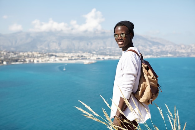 Attractive young Afro American hiker carrying small backpack contemplating amazing view of azure ocean, mountains and town below relaxing on top of rock after exhaustible climbing on sunny day