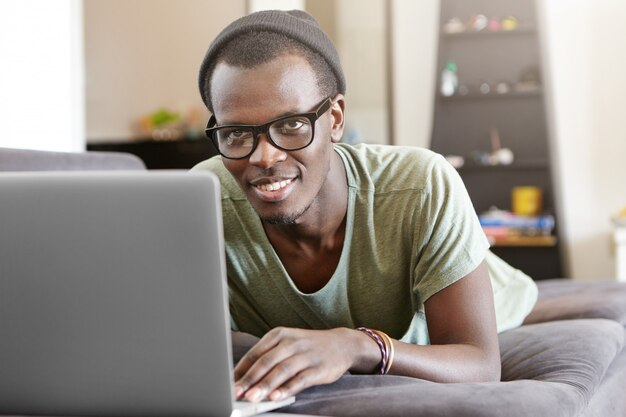 Attractive young Afro American male relaxing at home with notebook pc, lying on grey sofa alone after work. Black hipster having video call