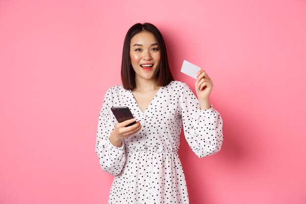 매력적인 젊은 아시아 여성이 신용카드를 들고 인터넷 구매를 하는 휴대전화를 온라인으로 주문합니다.