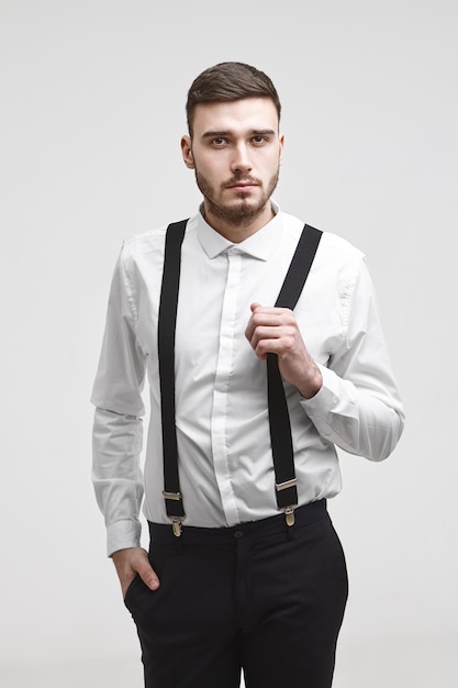 Attractive young bearded corporate worker wearing black trousers and white shirt posing isolated, holding hand in pocket, looking at camera with serious expression, pulling one strap of his suspenders