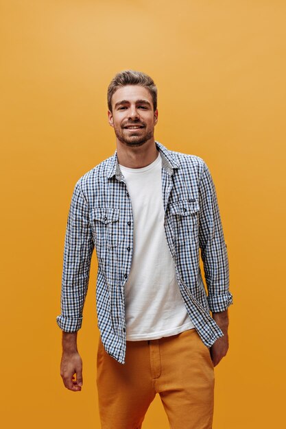 Attractive young bearded man in blue checkered shirt and white tshirt looks selfconfident and poses on orange background