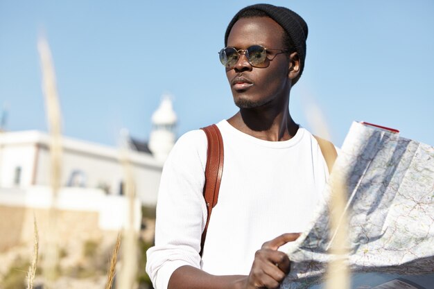 привлекательный молодой черный мужчина турист в модных солнечных очках и шляпе, держа бумажную карту и оглядываясь с серьезным сосредоточенным выражением, пытаясь найти дорогу в отель после того, как потерял