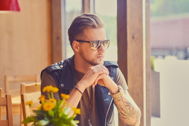 Attractive young blond bearded male dressed in a leather jacket sits at the table in a cafe.