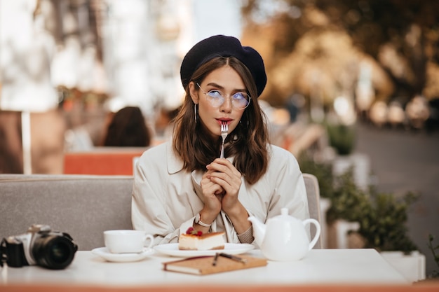 Attractive young brown-haired girl in glasses, vintage beret and beige trench coat, chilling at city cafe terrace, eating cheesecake and tea, thinking about something